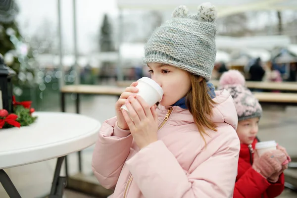Two adorable sisters drinking hot chocolate on traditional Christmas fair in Riga, Latvia. Children enjoying sweets, candies and gingerbread on Xmas market. — Stock Photo, Image