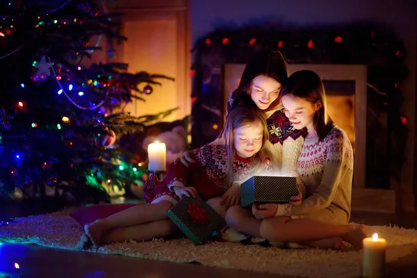 Happy young mother and her two small daughters opening a magical Christmas gift by a fireplace in a cozy dark living room on Christmas eve. — Stock Photo, Image