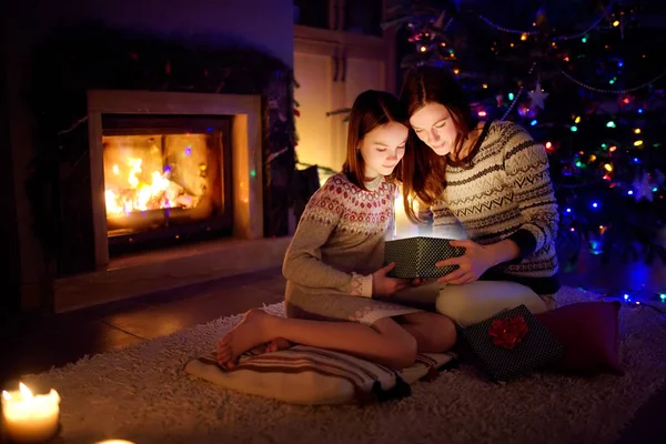 Happy young mother and her young daughter opening a magical Christmas gift by a fireplace in a cozy dark living room on Christmas eve. — Stock Photo, Image