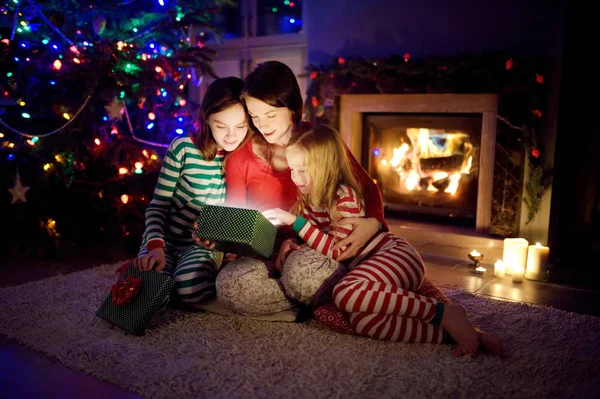 행복 한 젊은 어머니와 두 어린 딸 이 크리스마스 이브 때 아늑 한 거실에서 벽난로로 마법의 크리스마스 선물을 여는 모습. — 스톡 사진