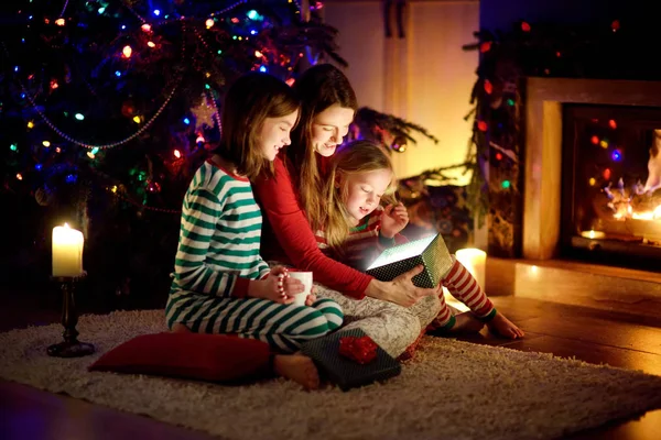 Szczęśliwa młoda matka i jej dwie małe córki otwierają magiczny prezent świąteczny przy kominku w przytulnym ciemnym salonie w Wigilię. — Zdjęcie stockowe