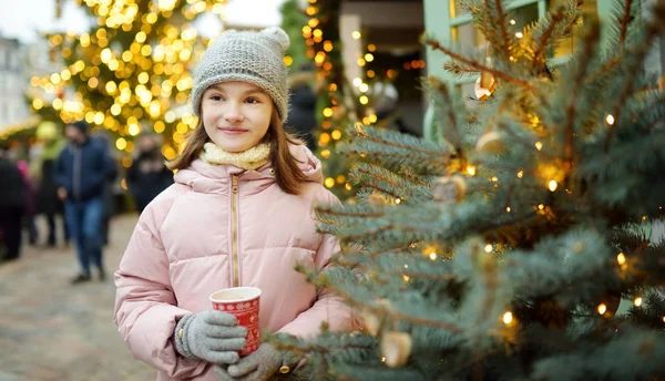 Menina bonito beber chocolate quente na feira de Natal tradicional em Riga, Letónia. Criança desfrutando de doces, doces e pão de gengibre no mercado de Natal . — Fotografia de Stock