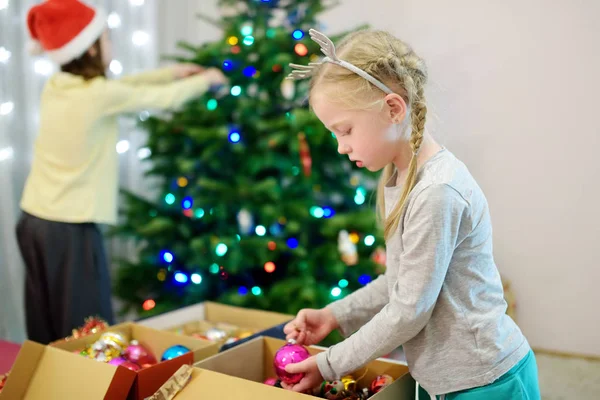 Két imádnivaló nővér, akik egy karácsonyfát díszítenek színes üvegfalatokkal otthon. Családi szabadidő a csodálatos karácsony idején. — Stock Fotó