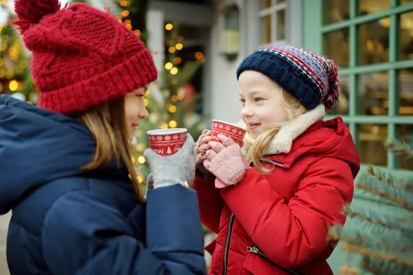 Duas irmãs adoráveis bebendo chocolate quente na tradicional feira de Natal em Riga, Letônia. Crianças desfrutando de doces, doces e pão de gengibre no mercado de Natal . — Fotografia de Stock