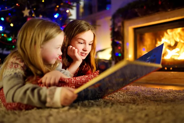 Jovens irmãs felizes lendo um livro de história juntas por uma lareira em uma acolhedora sala de estar escura na véspera de Natal. Celebrando o Natal em casa . — Fotografia de Stock