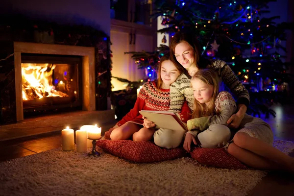 圣诞节前夕，妈妈和她的两个可爱的小女儿在温暖舒适的客厅里用平板电脑在家里壁炉边. — 图库照片