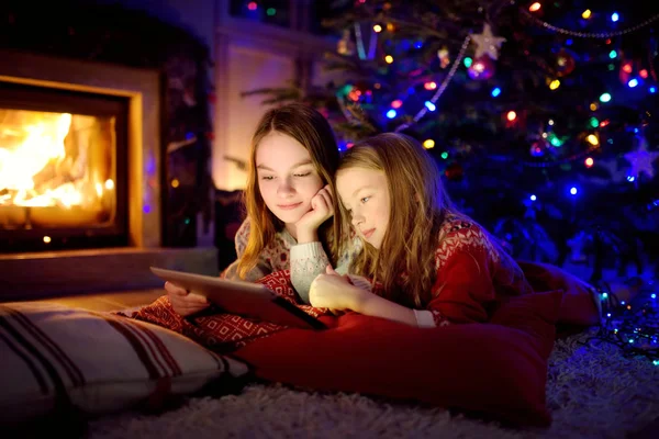 Two cute young sisters using a tablet pc at home by a fireplace in warm and cozy living room on Christmas eve. — Stock Photo, Image