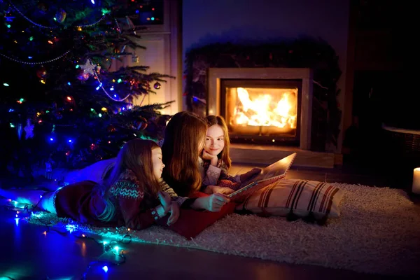 Feliz madre joven y sus hijas leyendo un libro de cuentos juntos junto a una chimenea en una acogedora sala de estar oscura en la víspera de Navidad. Celebrando la Navidad en casa . — Foto de Stock