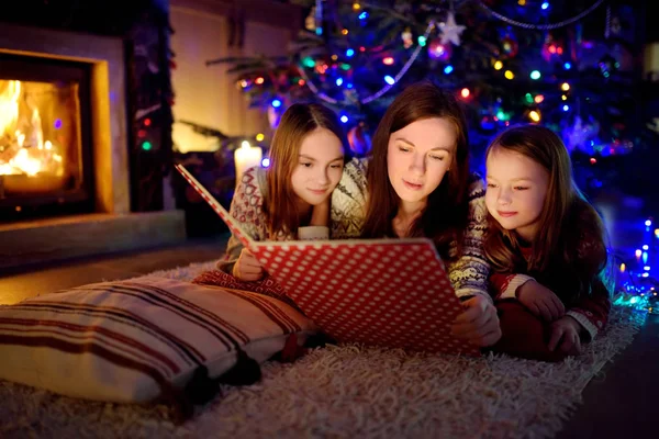 Jovem mãe feliz e suas filhas lendo um livro de história juntos por uma lareira em uma acolhedora sala de estar escura na véspera de Natal. Celebrando o Natal em casa . — Fotografia de Stock