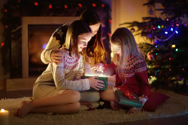 Šťastná mladá matka a její dvě malé dcery otevírají kouzelný vánoční dárek u krbu v útulném tmavém obývacím pokoji na Štědrý večer. — Stock fotografie