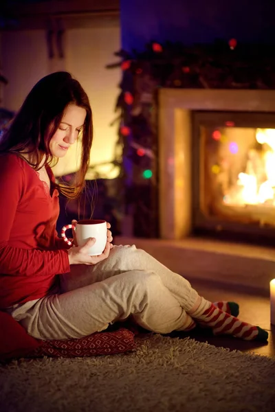 Happy young woman having a cup of hot chocolate by a fireplace in a cozy dark living room on Christmas eve. Celebrating Xmas at home.