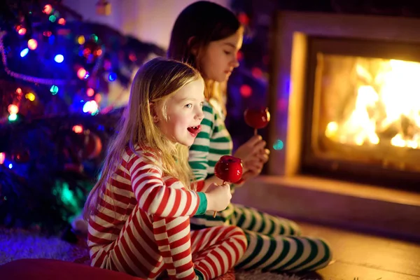 Hermanas jóvenes felices comiendo manzanas rojas cubiertas de azúcar juntos junto a una chimenea en una acogedora sala de estar oscura en la víspera de Navidad. Celebrando la Navidad en casa . —  Fotos de Stock