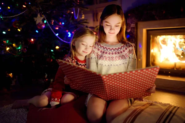 Glada unga systrar läser en sagobok tillsammans vid en öppen spis i ett mysigt mörkt vardagsrum på julafton. Fira jul hemma. — Stockfoto