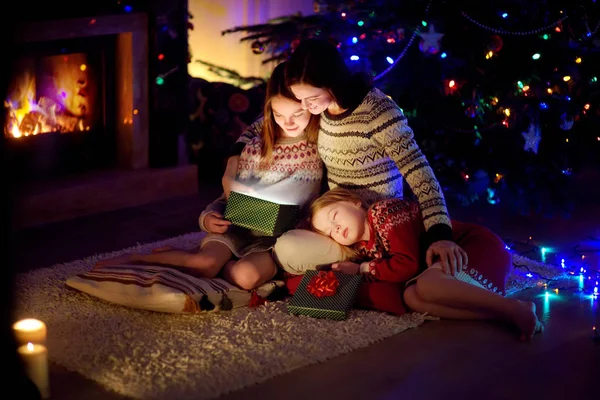 Happy young mother and her two small daughters opening a magical Christmas gift by a fireplace in a cozy dark living room on Christmas eve.