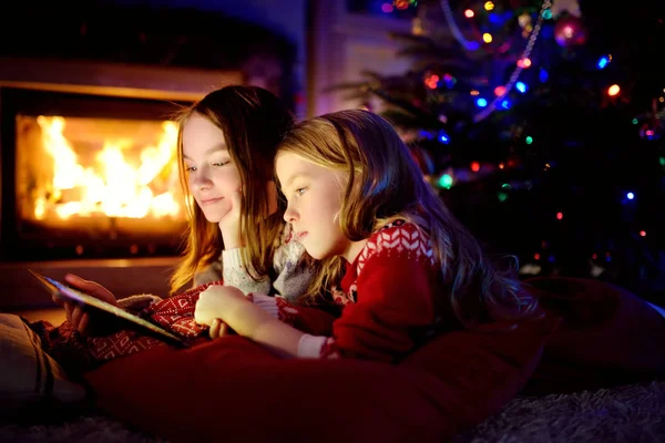 Two cute young sisters using a tablet pc at home by a fireplace in warm and cozy living room on Christmas eve. — Stock Photo, Image