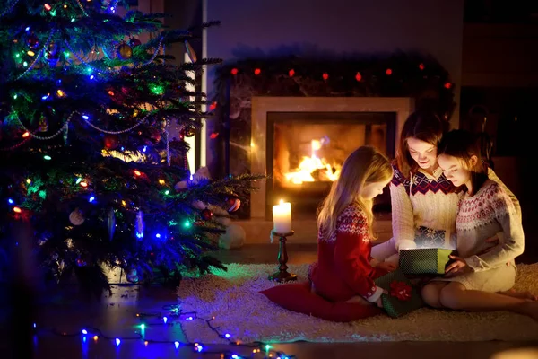 Feliz madre joven y sus dos hijas pequeñas abriendo un regalo mágico de Navidad junto a una chimenea en una acogedora sala de estar oscura en la víspera de Navidad . — Foto de Stock