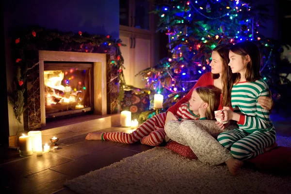 Happy young mother and her daughters having a good time sitting together by a fireplace in a cozy dark living room on Christmas eve. Celebrating Xmas at home. — Stock Photo, Image