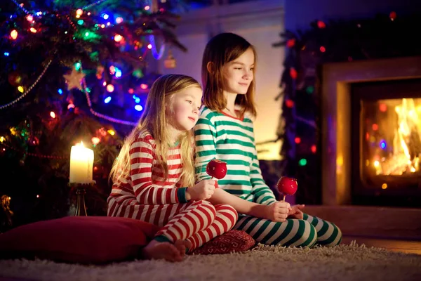 Irmãs felizes comendo maçãs vermelhas cobertas com açúcar gelado junto a uma lareira em uma acolhedora sala de estar escura na véspera de Natal. Celebrando o Natal em casa . — Fotografia de Stock