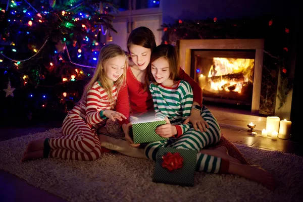 Ευτυχισμένη νεαρή μητέρα και οι δύο μικρές κόρες της ανοίγουν ένα μαγικό χριστουγεννιάτικο δώρο δίπλα σε ένα τζάκι σε ένα άνετο σκοτεινό σαλόνι την παραμονή των Χριστουγέννων. — Φωτογραφία Αρχείου