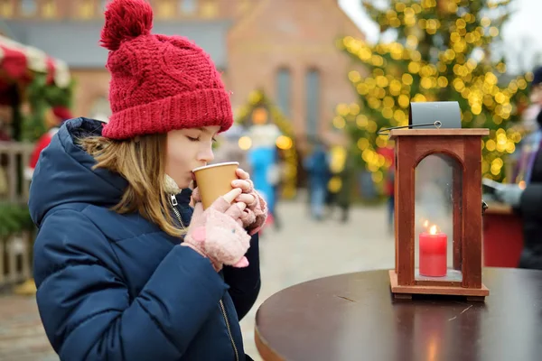 ラトビアのリガで開催される伝統的なクリスマスフェアでホットチョコレートを飲むかわいい女の子。クリスマスマーケットでお菓子、キャンディー、ジンジャーブレッドを楽しむ子供. — ストック写真