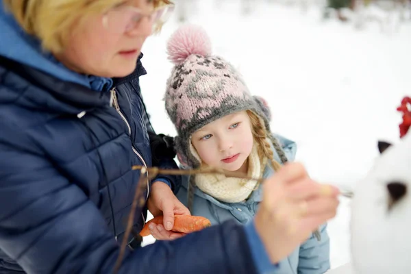 かわいい女の子と彼女の祖母は裏庭に雪だるまを構築します。雪の中で遊ぶかわいい子供. — ストック写真