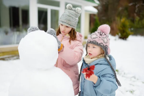 Adorable young girls building a snowman in the backyard. Cute children playing in a snow. Winter activities for kids. — Stock Photo, Image
