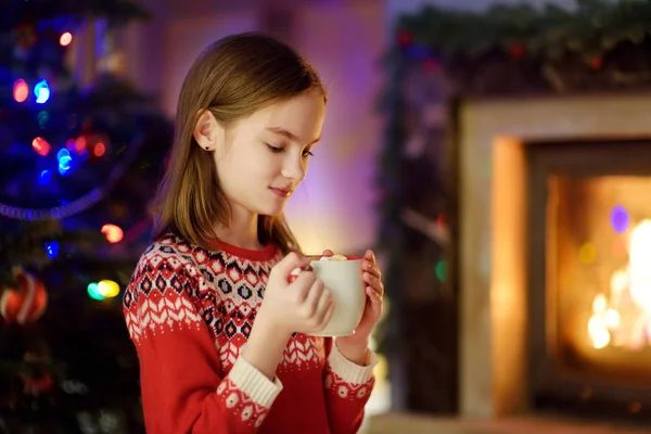 Happy young girl having a cup of hot chocolate by a fireplace in a cozy dark living room on Christmas eve. Celebrating Xmas at home. — Stock Photo, Image