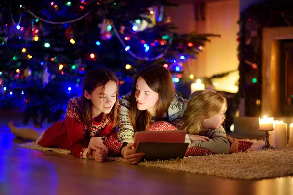 Mor och hennes två söta unga döttrar använder en surfplatta hemma vid en öppen spis i varmt och mysigt vardagsrum på julafton. — Stockfoto