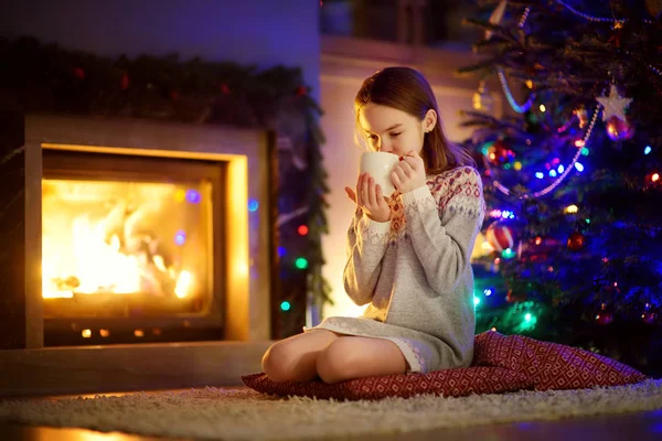 Joyeux jeune fille ayant une tasse de chocolat chaud près d'une cheminée dans un salon sombre confortable la veille de Noël. Célébrer Noël à la maison . — Photo