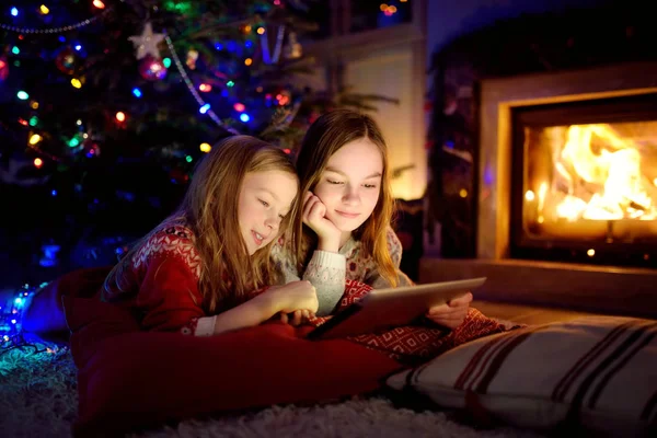 Two cute young sisters using a tablet pc at home by a fireplace in warm and cozy living room on Christmas eve. — Stock Photo, Image