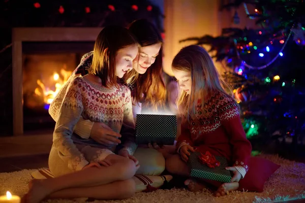Boldog fiatal anya és két kislánya nyit egy varázslatos karácsonyi ajándékot egy kandalló egy hangulatos sötét nappaliban karácsony este. — Stock Fotó