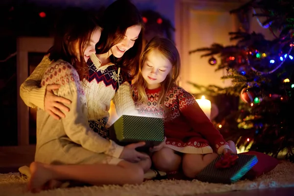 Happy young mother and her two small daughters opening a magical Christmas gift by a fireplace in a cozy dark living room on Christmas eve. — Stock Photo, Image