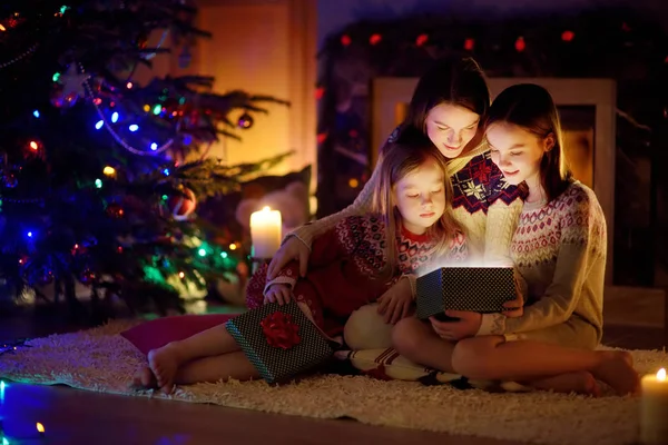 Šťastná mladá matka a její dvě malé dcery otevírají kouzelný vánoční dárek u krbu v útulném tmavém obývacím pokoji na Štědrý večer. — Stock fotografie