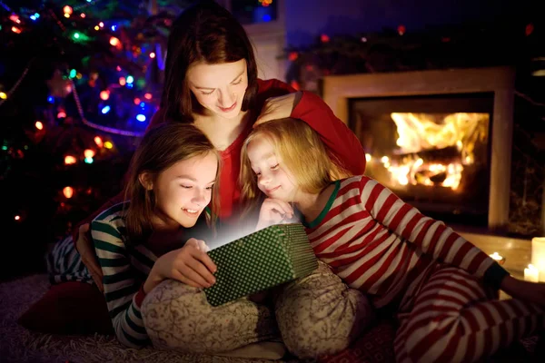 Happy young mother and her two small daughters opening a magical Christmas gift by a fireplace in a cozy dark living room on Christmas eve.