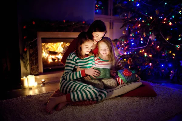 Happy young mother and her two small daughters opening a magical Christmas gift by a fireplace in a cozy dark living room on Christmas eve. — Stock Photo, Image