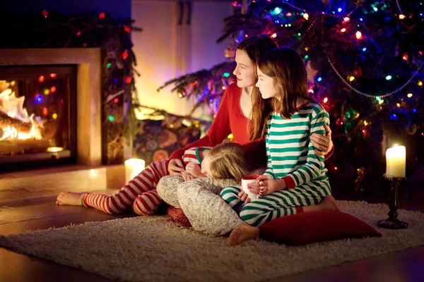 Glad ung mor och hennes döttrar har det trevligt att sitta tillsammans vid en öppen spis i ett mysigt mörkt vardagsrum på julafton. Fira jul hemma. — Stockfoto
