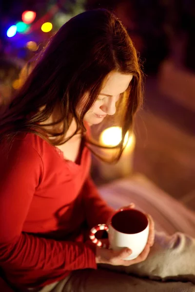 Vrolijke jonge vrouw met een kopje warme chocolademelk bij een open haard in een gezellige donkere woonkamer op kerstavond. Thuis Kerstmis vieren. — Stockfoto