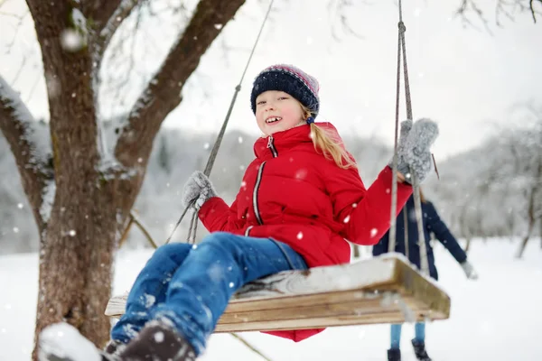 Two adorable young girls having fun on a swing together in beautiful winter park. Cute sisters playing in a snow. — Stock Photo, Image