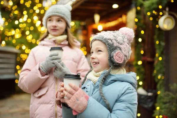 Deux adorables sœurs boivent du chocolat chaud lors de la foire traditionnelle de Noël à Riga, en Lettonie. Enfants dégustant des bonbons, des bonbons et du pain d'épice sur le marché de Noël . — Photo