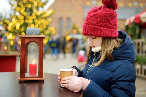 Милая молодая девушка пьет горячий шоколад на традиционной рождественской ярмарке в Риге, Латвия. Ребенок наслаждается сладостями, сладостями и пряниками на рождественском рынке . — стоковое фото