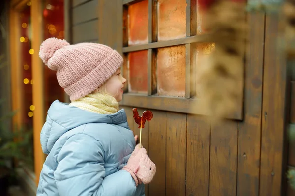 Menina bonito ter pirulito em forma de galo na feira de Natal tradicional em Riga, Letônia. Criança desfrutando de doces, doces e pão de gengibre no mercado de Natal . — Fotografia de Stock