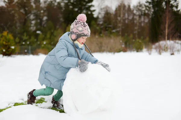 Suloinen nuori tyttö rakentamassa lumiukkoa takapihalle. Söpö lapsi leikkii lumessa . — kuvapankkivalokuva