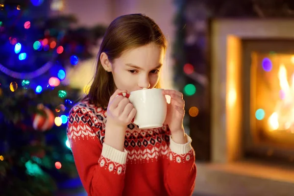 Счастливая молодая девушка с чашкой горячего шоколада у камина в уютной темной гостиной в канун Рождества. Празднование Рождества дома . — стоковое фото