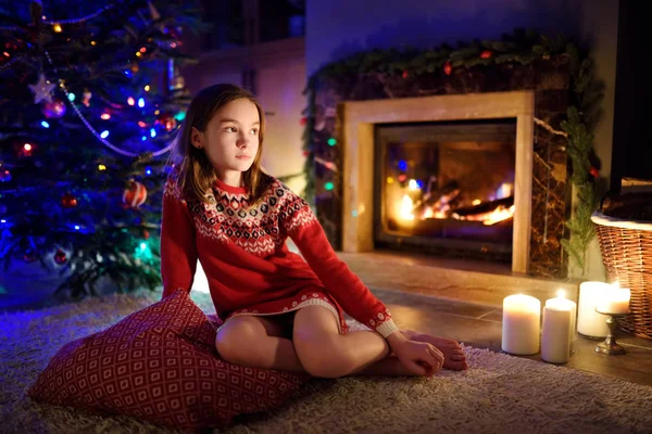 Chica joven feliz pasar un buen rato sentado junto a una chimenea en una acogedora sala de estar oscura en la víspera de Navidad. Celebrando la Navidad en casa . — Foto de Stock