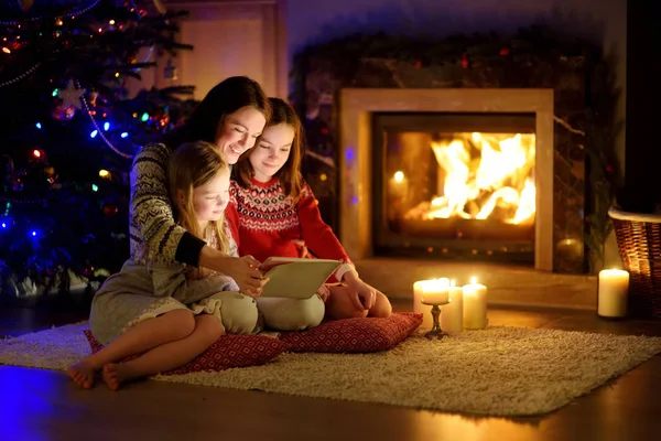 Mother and her two cute young daughters using a tablet pc at home by a fireplace in warm and cozy living room on Christmas eve. — Stock Photo, Image