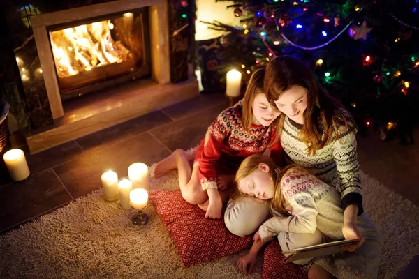 Mother and her two cute young daughters using a tablet pc at home by a fireplace in warm and cozy living room on Christmas eve. — Stock Photo, Image