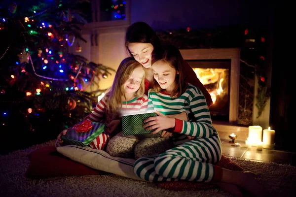 행복 한 젊은 어머니와 두 어린 딸 이 크리스마스 이브 때 아늑 한 거실에서 벽난로로 마법의 크리스마스 선물을 여는 모습. — 스톡 사진