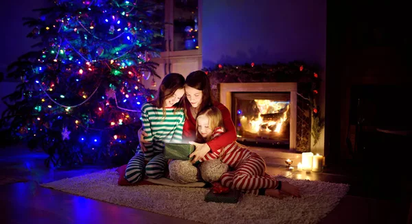 Happy young mother and her two small daughters opening a magical Christmas gift by a fireplace in a cozy dark living room on Christmas eve.