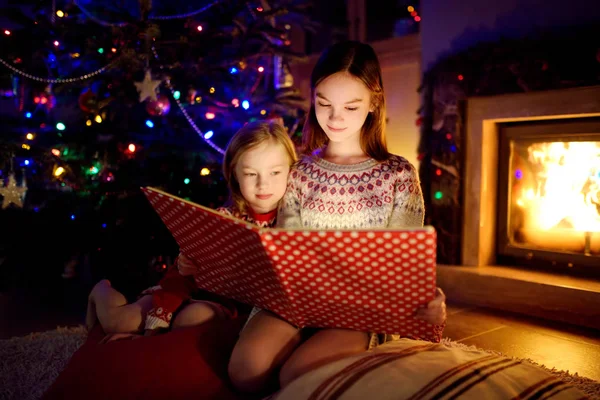Jovens irmãs felizes lendo um livro de história juntas por uma lareira em uma acolhedora sala de estar escura na véspera de Natal. Celebrando o Natal em casa . — Fotografia de Stock