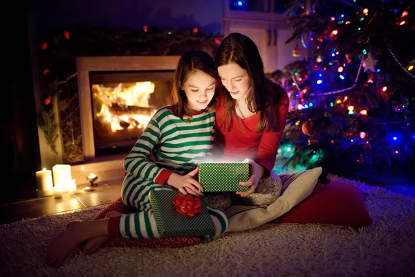 Gelukkig jong moeder en haar jonge dochter openen een magisch kerstcadeau bij een open haard in een gezellige donkere woonkamer op kerstavond. — Stockfoto